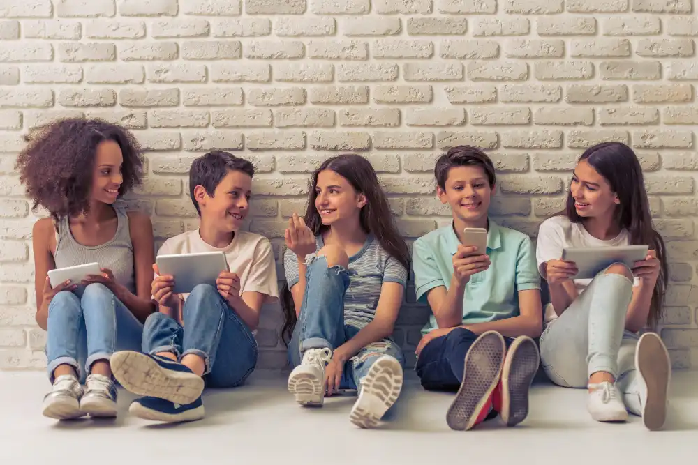 Group of young boys and girls using devices, talking and smiling, sitting against white brick wall